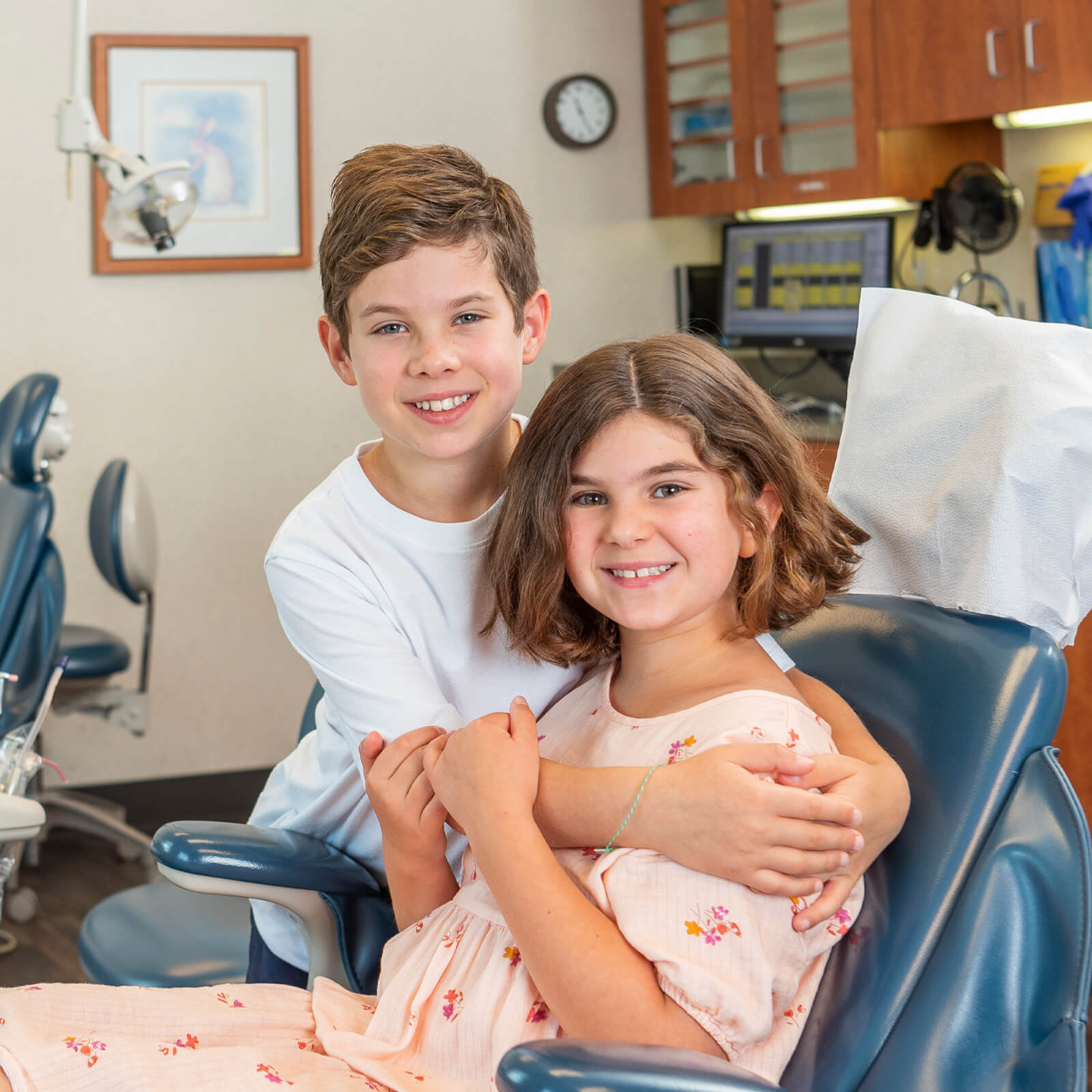 Brother hugging his sister who is sitting in a dentist chair