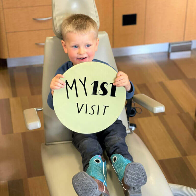 Toddler in a dentist chair holding a sign that reads 